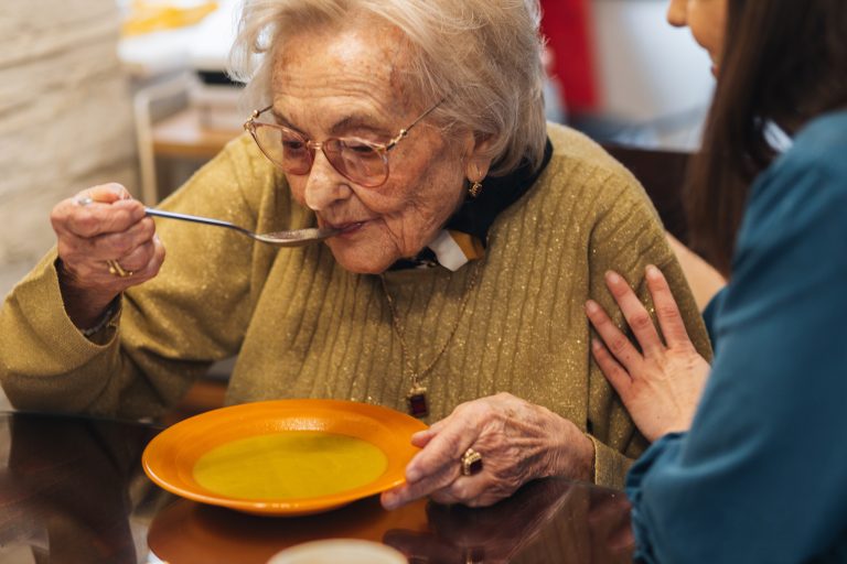 Elderly woman eating soup with a young woman sitting beside her.